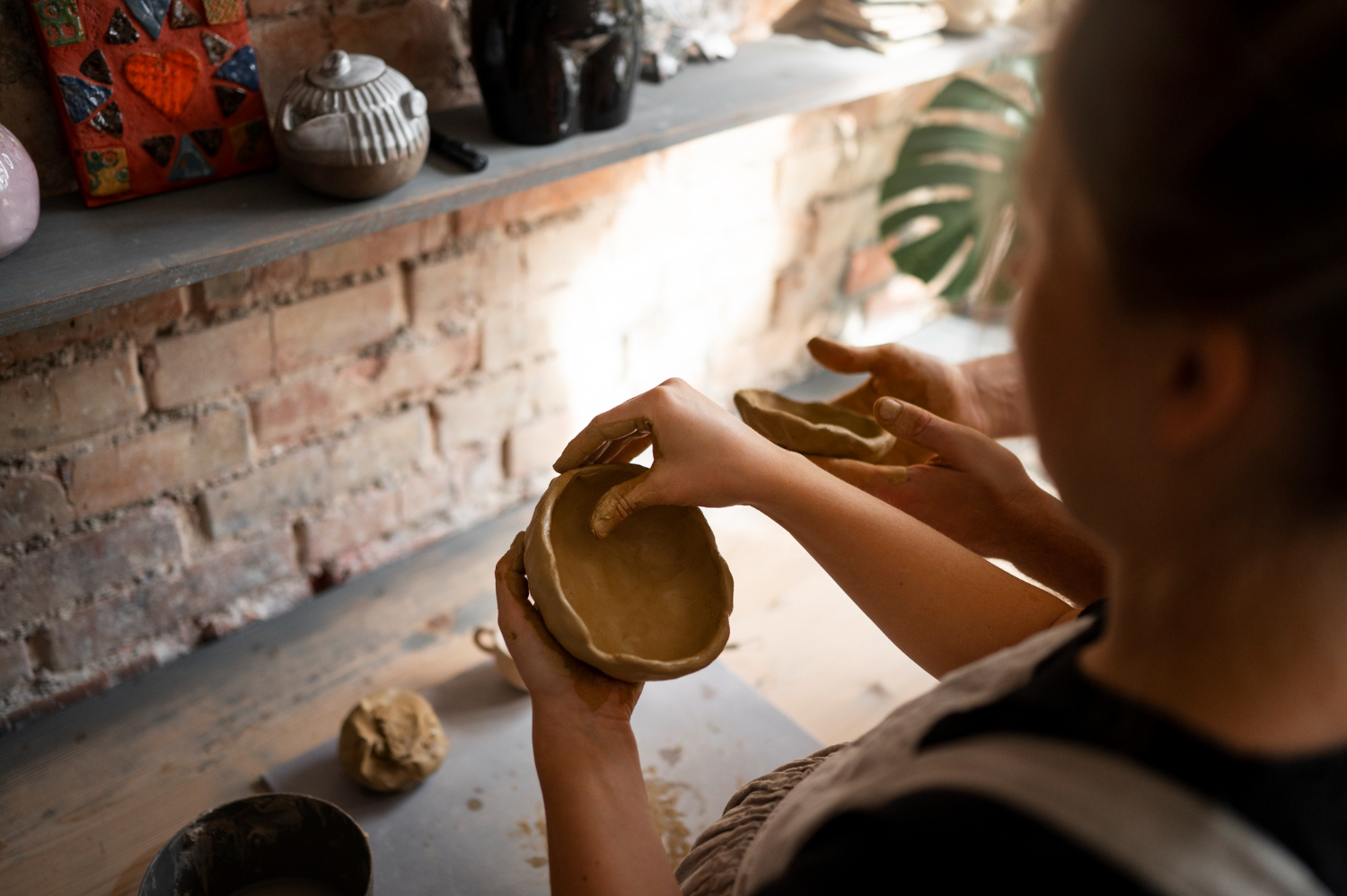 Two people shaping a hand-built clay bowl together at the studio's ceramics table